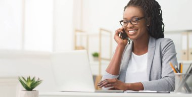 Patroniçe. Modern ofiste dizüstü bilgisayarla çalışırken cep telefonuyla konuşan Afro İş kadını, Geniş Yatay Sancak, Boş Alan Panorama