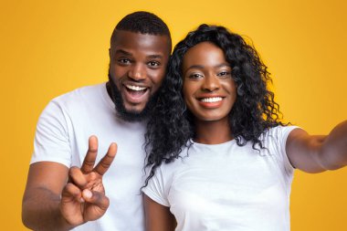 Bir çift fotoğraf için poz veriyor, gülümsüyor ve kameraya bakıyor. Kadın kolunu uzatıp fotoğraf çekerken, adam barış işareti gösteriyor..