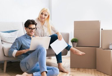 Bir çift yeni evlerinin zemininde oturuyor, adam dizüstü bilgisayar kullanıyor, kadın ise bir belge tutuyor..