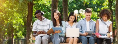 Eğitim konsepti. Laptop ve tablet bilgisayarlı bir grup üniversite öğrencisi kampüste birlikte çalışıyor, dışarıda bankta oturuyor.