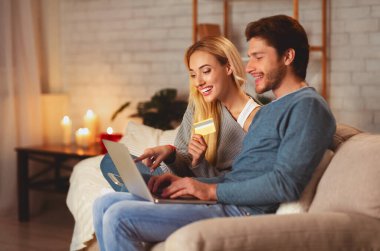 İnternetten alışveriş. Mutlu eşler dizüstü bilgisayar ve kredi kartı tutuyor. Evdeki koltukta oturan internet mağazasından bir şeyler sipariş ediyorlar.