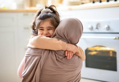 Anneler Günü Konsepti. Neşeli küçük kız tesettürdeki Müslüman annesine sımsıkı sarılıyor, İslami kadın gülümseyen çocuğu kucaklıyor, mutlu aile mutfaklarında birbirine bağlanıyor,