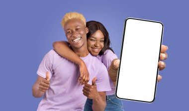 Siyahi bir çift, her ikisi de gülümsüyor, mavi arka planda birlikte poz veriyorlar. Kadın elinde beyaz, boş ekranlı bir akıllı telefon tutuyor ve işaret parmağıyla işaret ediyor. Adam başparmağını kaldırıyor..