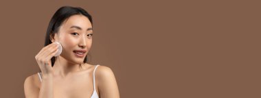 Şehvetli Asyalı kadın elinde pamuk tutuyordu, kahverengi stüdyo arka planında toner ile yüzünü temizliyordu, kameraya bakıp gülümsüyordu. Mutlu kadın taze nemlendirilmiş yüz derisinden zevk alıyor, kopyalama alanı