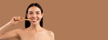 Çekici, neşeli esmer, uzun saçlı, çıplak kadın. Dişlerini organik ahşap diş fırçasıyla fırçalıyor. Bej rengi stüdyo arka planında izole edilmiş dişleri temizliyor.