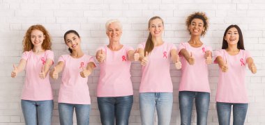 Göğüs kanseri tedavisi. Farkındalık Tişörtleri ve Pembe Kurdeleli Mutlu Karışık Kadınlar Beyaz Duvar 'ın Üzerinde Başparmak Kalkıyor