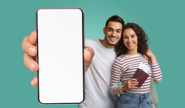 Orta doğulu bir çift turkuaz bir arka fonun önünde, kameraya doğru boş ekranlı bir akıllı telefon tutarak yan yana duruyorlar. Kadının elinde pasaportu ve uçak biletleri var..