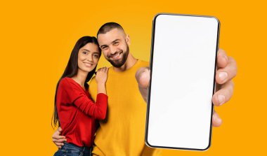 Mutlu genç bir çift, sarı bir arka plan önünde kameraya gülümseyerek yan yana durur. Adam önünde beyaz bir ekranla akıllı bir telefon tutuyor..