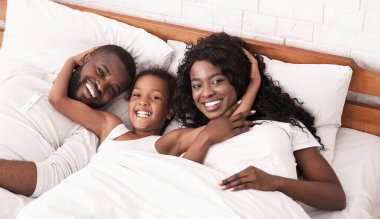 Mutlu Afro-Amerikan ebeveynler ve kızları yatakta birlikte rahatlıyor ve kameraya bakıyor.