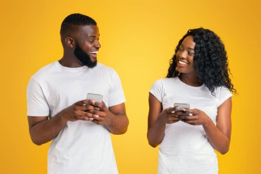 Beyaz tişörtler giymiş gülümseyen siyah bir çift, sarı arka planda birbirlerine bakarken akıllı telefonlar tutuyorlar..