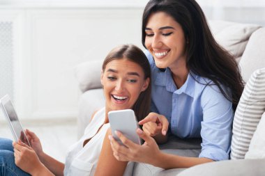 Aletler. Telefonunu ve dijital tabletini kullanan gülümseyen iki kız. Kapalı koltukta otururken çekilmiş resimlerini gösteriyorlar.