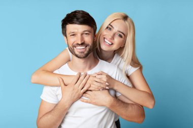 Uzun, sarı saçlı bir kadın, bir erkeği arkadan kucaklıyor. Her ikisi de kameraya parlak bir şekilde gülümsüyor. Basit beyaz tişörtler giyiyorlar ve birbirlerine eşlik etmekten zevk alıyor gibi görünüyorlar.
