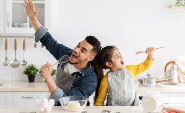 Neşeli Arap baba ve küçük kızı mutfakta birlikte yemek pişirirken eğleniyorlar. Mutlu ortadoğulu baba ve çocuk şarkı söyleyip eğleniyorlar. Spatula ve çırpma aletlerini mikrofon olarak kullanıyorlar.