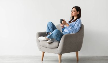 Rahat giyinen genç bir bayan bacak bacak üstüne atmış bir şekilde koltukta oturuyor, sıcak bir fincan kahvenin üzerinde ellerini ısıtıyor, beyaz stüdyo duvarına yaslanmış sıcak içkinin keyfini çıkarıyor,