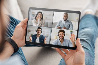 Adam elinde bir tablet tutuyor ve dört kişiyle online video konferansına katılıyor, evdeki kanepede dinleniyor.