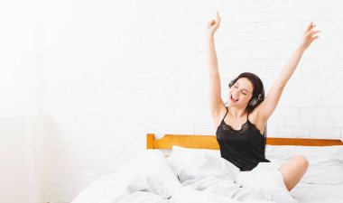 Genç bir kadın beyaz bir yatağa oturmuş kollarını kaldırarak kulaklıkla müzik dinliyor. Gerçek bir mutluluk ve rahatlama anında gibi görünüyor, kopyalama alanı.