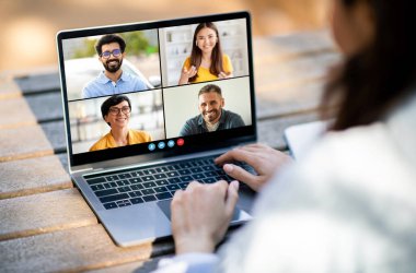 Bir kadın ahşap bir yüzeyde oturuyor ve bir video konferansına katılıyor. Dizüstü bilgisayarı dört katılımcıyı ayrı video çerçevelerinde gösteren bir ekrana açık