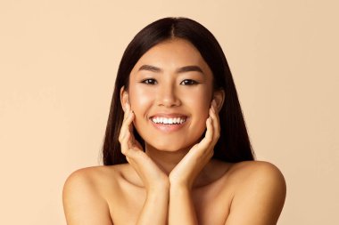 Uzun siyah saçlı genç bir Asyalı kadın kameraya sıcak bir şekilde gülümsüyor. Makyaj yapmamış ve cildi temiz. Elleri yavaşça yanaklarında dinleniyor.