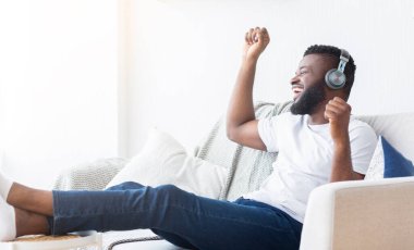 Kulaklık takan Afrikalı Amerikalı bir adam bir kanepede oturuyor ve müzik dinliyor. Gülümsüyor ve elini havaya kaldırıyor, sanki dans ediyormuş ya da şarkı söylüyormuş gibi..