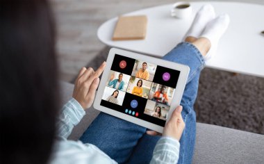 Bir kadın elinde tablet tutarken, ayakları sehpanın üzerinde dinleniyor. Ekranda birden fazla katılımcının katıldığı bir video konferansı gösteriliyor.