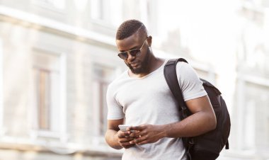 Güneş gözlüğü takan genç bir adam şehirde yürürken akıllı telefonunu kullanır..