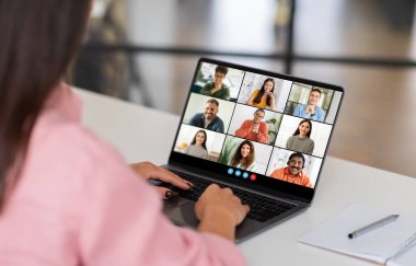 Ev ofisinin masasında oturan bir kadın grup video konferansına katılırken dizüstü bilgisayarına bakıyor. Ekranda birkaç katılımcı ızgara biçimi, webinar görüntüler