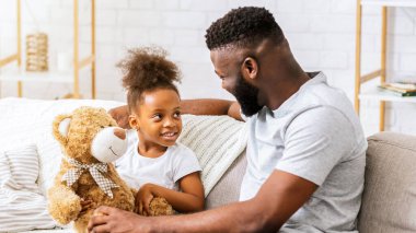 Bir baba ve kızı, her ikisi de Afrikalı Amerikalı, bir kanepede birlikte rahatlatıcı bir anın tadını çıkarıyorlar. Kızı oyuncak ayıyı tutuyor ve babasına gülümsüyor. O da gülümsüyor..