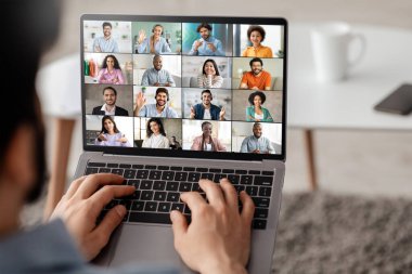 Bir adam kanepede oturuyor ve video konferansına katılmak için dizüstü bilgisayar kullanıyor. Dizüstü bilgisayarın ekranında birkaç farklı insanı gösteren bir video yayını var.