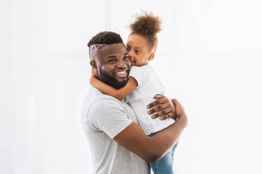 Siyah baba kızını kollarında tutarken gülümsüyor. Kız ona sıkıca sarılıyor ve başını omzuna yaslıyor. Beyaz bir odada duruyorlar.