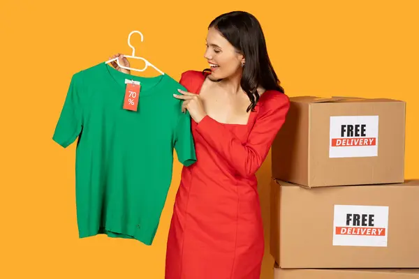 stock image A woman in a red dress stands in front of a bright yellow background holding a green t-shirt with a 70 off tag on a hanger. Behind her are two stacked cardboard boxes labeled Free Delivery.