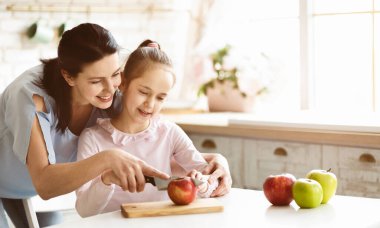 Bir anne ve küçük kızı mutfakta birlikte sağlıklı bir aperatif hazırlıyorlar. Anne bir bıçak tutuyor ve kızı dikkatlice izlerken bir elmayı kesiyor..