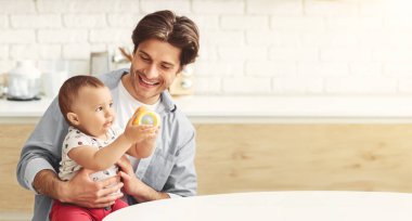 Gülümseyen bir baba, bebeğini mutfak ortamında kollarına alır. Bebek babasının tuttuğu bir oyuncağa uzanıyor. Baba oğluna sevgi ve sevgiyle bakıyor. Kopya uzayı.