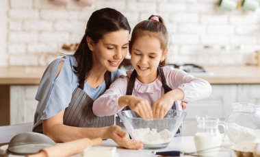Bir anne ve genç kızı, aydınlık ve modern bir mutfakta birlikte yemek pişiriyorlar. Kız cam bir kaseye un eklerken, anne gülümseyerek bakıyor..