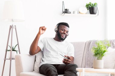 Beyaz tişörtlü Afro-Amerikan bir adam oturma odasında bir kanepede oturuyor. Elinde akıllı bir telefonla gülümsüyor ve kulaklıkla müzik dinliyor.