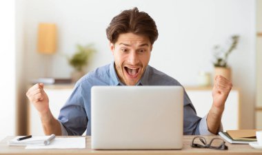 Bir adam masada oturmuş, dizüstü bilgisayarına heyecanlı bir ifadeyle bakıyor ve yumruklarını kaldırıyor..