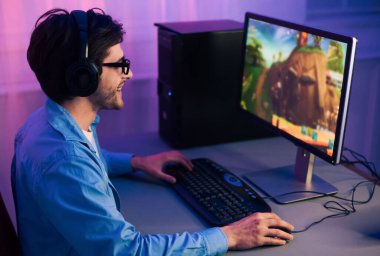 Genç bir adam bilgisayarının başına oturmuş kulaklık takıyor ve video oyunu oynuyor. O gülümsüyor ve eğleniyor gibi görünüyor. Oyun bilgisayar ekranında ve adamda gösterilir