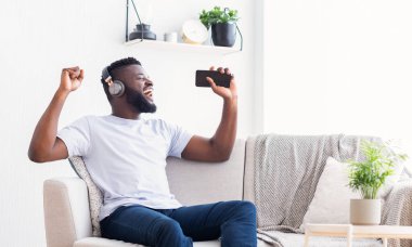 Genç bir siyah adam oturma odasında beyaz bir kanepede oturuyor, kulaklık takıyor ve akıllı telefon tutuyor. Müziğe eşlik ediyor, yüzü neşeyle aydınlanıyor, kopyalama alanı.