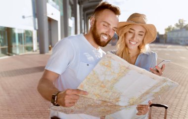 Turizm ve Seyahat Konsepti. İki sevimli turist ellerinde kağıt harita ve havaalanı yakınlarında açık havada cep telefonu kullanıyorlar.