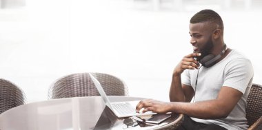 Bir adam dışarıda bir kafede oturuyor ve laptopuyla çalışıyor. Boynunda kulaklık var ve gülümsüyor..