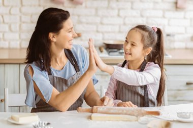 Bir anne ve kızı mutfakta beraberler, önlük giyiyorlar ve birbirlerine beşlik çakıyorlar. Gülümsüyorlar ve yemek pişirirken eğleniyormuş gibi görünüyorlar..