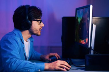 Gözlüklü ve kulaklık takan genç bir adam bilgisayarının başına oturmuş ve bir video oyunu oynamaya odaklanmış. Odası loş bir şekilde mavi ve mor renklerle aydınlatılmış, atmosferik bir ortam yaratıyor..