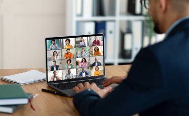 Önünde bir dizüstü bilgisayar olan bir işadamı masada oturuyor ve her karede birçok insanın olduğu bir video konferans ekranı gösteriyor. Bilgisayarını online bir toplantı için kullanıyor..