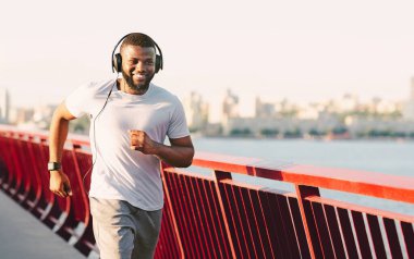 Arka planda şehir silüeti olan bir köprüde koşarken genç bir adam gülümsüyor. Kulaklık ve beyaz bir tişört giyiyor..