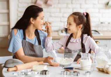 Bir anne ve kız mutfakta birlikte yemek pişirirken eğleniyorlar. Genç kız, her ikisi de birbirlerine gülümserken annesinin burnuna neşeyle dokunuyor..