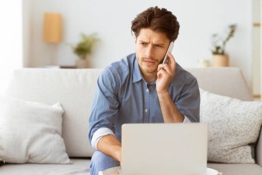 Genç bir adam kanepede oturur, telefonuyla konuşurken dizüstü bilgisayarıyla çalışır. Bir sohbete odaklanmış gibi görünüyor..