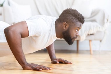 Beyaz tişörtlü Afro-Amerikan bir adam evinde ahşap zeminde şınav çekerken görülüyor. Egzersize odaklanmış ve güçlü ve kararlı görünüyor..