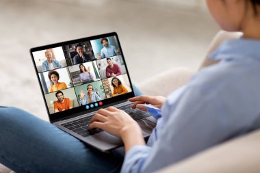 Görüntü, kanepede oturan bir kadının dizüstü bilgisayardaki bir video konferansına katıldığını gösteriyor. Dizüstü bilgisayarı açık ve her birinde farklı bir kişinin yer aldığı dokuz video yayınını gösteriyor..