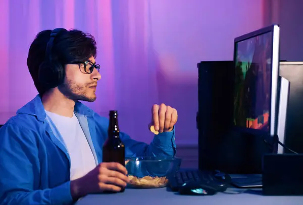 stock image This image shows a young man playing video games while wearing headphones. He is sitting in front of a computer with a controller in his hand. He is enjoying snacks and a beer while he plays.