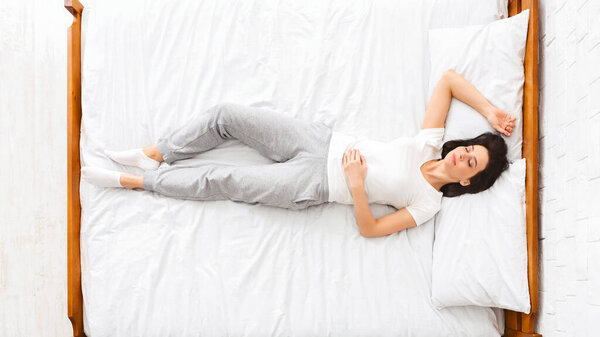 Вид сверху женщины, лежащей на кровати, покрытой хрустящими белыми простынями. Она отдыхает комфортно, ее глаза закрыты в момент релаксации.