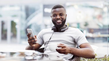 Bir adam akıllı telefonunu kullanıp açık bir kafede kahve içerken gülümsüyor..